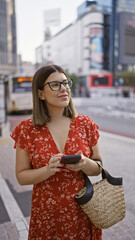 Canvas Print - Smiling beautiful hispanic woman in glasses uses phone on bustling tokyo street, enjoying modern city's vibe, cheerfully texting away