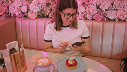 Woman checks phone in pink cafe with dessert, coffee, flowers, glasses, youthful, brunette, seated, technology