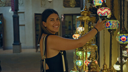 Wall Mural - Young brunette woman smiling while exploring traditional lamp souk in dubai, expressing joy and curiosity.