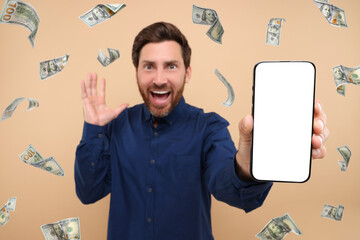 Happy man showing mobile phone with empty screen under money rain on dark beige background, selective focus