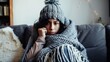 © VK Studio - A woman bundled in a cozy grey knit blanket and hat sits on a couch, looking thoughtful and warm in her home during a chilly day.