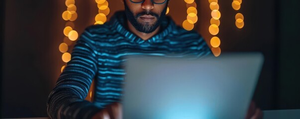 Focused man at laptop with hooded sweatshirt, working late. Blurred computer screen, bokeh lights in background, evening ambiance.