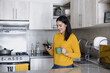 © Marcos - Latin adult woman drinking a coffee cup and using mobile phone in kitchen in Mexico Latin America, hispanic female at home