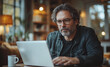© gabriele - A middle-aged man is sitting at a table in his bright living room, working on an open laptop computer with glasses and wearing casual . The background features white walls, large windows, green plant
