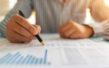 Poster - Close up of a professional analyzing financial documents with a pen, focusing on charts and graphs in a bright office setting.