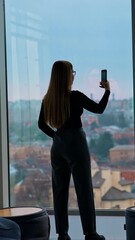 Sticker - Long-haired girl with a phone indoors. Young woman shooting a video of a beautiful evening view of a city landscape from the office window. Rear view. Vertical video