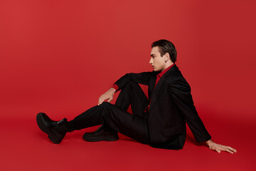 A man in a black suit and red shirt poses casually on a bright red backdrop.