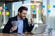 © Liubomir - Successful businessman celebrating financial achievement at office desk holding cash and working on laptop. Smiling man expressing joy and excitement at work environment.