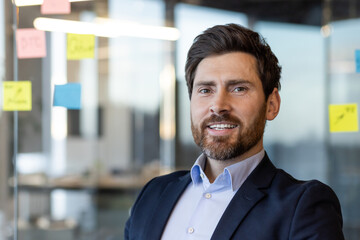 Wall Mural - Confident businessman in professional attire smiling in office setting with sticky notes on glass wall background. Concept of leadership, success, entrepreneurship.