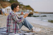 © ViDi Studio - Full body side view young fun woman wear shirt casual clothes hold in hand use mobile cell phone sitting rest on sea ocean sand shore beach outdoor seaside in summer day free time. Lifestyle concept