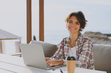 Wall Mural - Young smiling confident fun IT woman wear shirt casual clothes sitting alone at street cafe table in coffee shop working or studying on laptop pc computer rest in restaurant during free time outdoor.