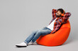 © ViDi Studio - Full body young happy smiling Indian man he wearing red shirt casual clothes sit in bag chair listen to music in headphones isolated on plain grey color background studio portrait. Lifestyle concept.