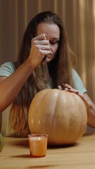 Wall Mural - Vertical video. In the Warm Light of Lamps at Home in the Evening, a Woman Carves a Jack-O'-Lantern from a Pumpkin, Forcefully Stabbing the Top with a Knife. Festive Preparations.