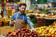 © Liubomir - Male grocer wearing apron arranging apples in produce section of grocery store. Focus on fresh fruit display with vibrant colors. Concept of retail, grocery, and healthy food shopping.