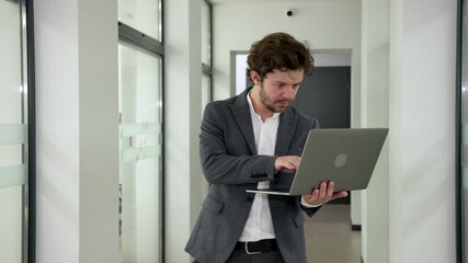 Confident young programmer or analyst walking through a modern office corridor, using a laptop. Perfect for showcasing tech skills and a dynamic work environment.