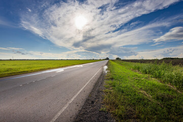 Wall Mural - A tranquil road winds through vibrant green fields, illuminated by sunlight breaking through a stunning, dynamic cloudscape overhead.