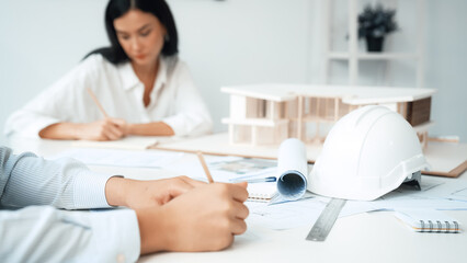 Wall Mural - Professional architect engineer drawing blueprint and working together with young beautiful caucasian coworker on meeting table with safety helmet, house model, blueprint scatter around. Immaculate.