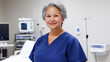 © wernerimages - A smiling nurse with short gray hair, wearing blue scrubs, standing in a medical facility with various medical equipment in the background.