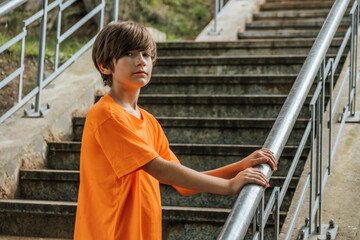 A young boy in an orange shirt and red shorts squats on stone steps, wearing black sneakers. His relaxed pose and casual clothing create a laid-back atmosphere. The background features worn stone step