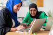 © Studio Marmellata - Two women in hijabs collaborate and smile while working on laptops at a desk in a modern office. The environment is bright and professional, reflecting teamwork and productivity