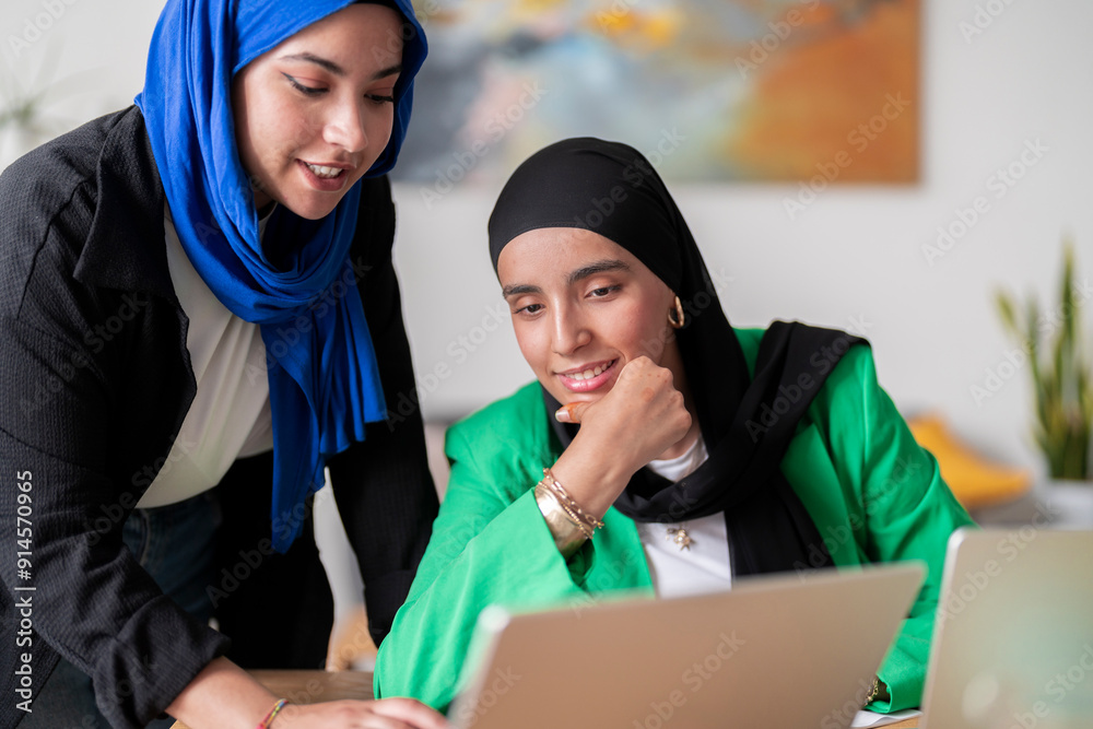 Two women in hijabs collaborate while looking at a laptop screen ...