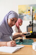 © Studio Marmellata - Two women in hijabs work in a 3D printing lab, one taking measurements while the other focuses on her laptop. The scene captures a blend of digital and physical workspaces.