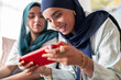 © Studio Marmellata - Two women in hijabs smile as they play a game on a tablet. Their expressions show joy and engagement, highlighting a fun and relaxed atmosphere with modern technology.