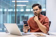 © Liubomir - Young professional using phone while working on laptop at office desk. Businessman wearing glasses and shirt focused on smartphone. Concept of multitasking, technology, modern work environment.