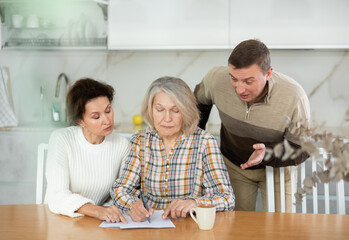 Competitive middle-aged man and woman conflicting and old woman writing inheritance sitting at kitchen table