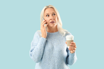 Wall Mural - Mature woman with takeaway cup of coffee talking by mobile phone on blue background