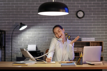 Canvas Print - Young woman sitting at workplace with modern laptop and talking with phone in office