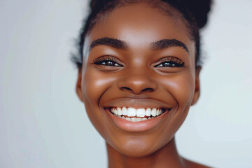 A closeup portrait of a young woman with dark skin smiling broadly, revealing her bright white teeth. Her eyes are open and sparkling, and her hair is styled in a bun.