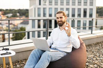 Wall Mural - Happy man working remotely on rooftop terrace using laptop giving okay sign. Comfortable workspace outdoors with cityscape in background. Remote work, freelance lifestyle, outdoor office.