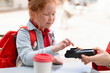 © Iryna - Young Girl Using  Payment Device in  Café, Wearing  Red Backpack and Denim Jacket, During a Sunny Day