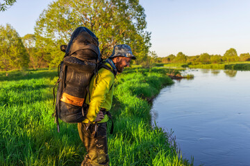 Wall Mural - A determined hiker stands by a tranquil river, lost in thought, surrounded by vibrant grass and trees as the sun rises in the early morning sky.