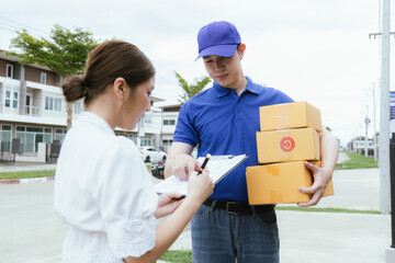 A cheerful Asian woman receives a package from a delivery man in uniform at her doorstep. The courier hands her a paper bag as she signs a receipt using her mobile, completing the home delivery servic