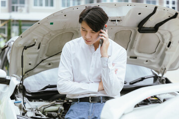 A bearded man in his 30s stands beside his broken-down car with the hood open, looking frustrated. Stranded on the roadside during the day, he uses his smartphone to seek insurance assistance for the 