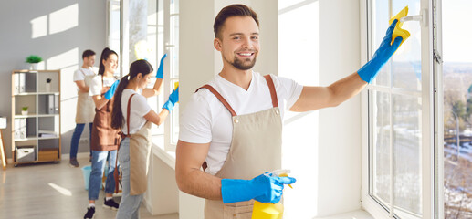 Wall Mural - Portrait of a young man from professional cleaning service using a duster and a spray, looking at camera and smiling while cleaning windows in the house with a team of janitors in background.