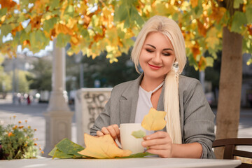 Wall Mural - A blonde woman sits at a table with a cup of coffee and a leaf on it. The scene is set in a city with a tree in the background.