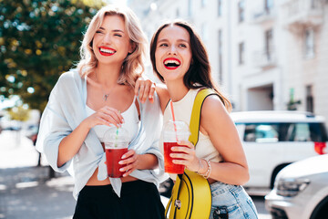 Two young beautiful smiling glam female in trendy summer clothes. Sexy women posing outdoors. Positive models holding and drinking fresh cocktail smoothie drink in plastic cup. Bright makeup, red lips