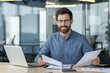 © Tetiana - Portrait of a smiling and successful young businessman sitting in the office at the table, holding documents and papers for a deal in his hands, confidently looking at the camera