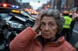 © Milos - An elderly woman stands distraught in front of a damaged car on a busy city street with emergency vehicles and personnel in the background during the day.