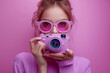 © Kmikhidov - A young woman wearing stylish pink sunglasses holds a camera close as she poses confidently against a bold purple background, capturing a moment of creativity and self-expression