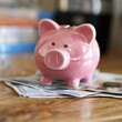 © Suritong - A cute pink piggy bank sitting on top of cash, symbolizing savings and financial planning at home.