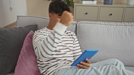 Canvas Print - Young hispanic man using tablet in modern living room, showing focused and happy expressions, wearing striped shirt, seated on comfortable sofa with cushions, enjoying his time indoors.