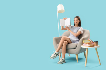 Wall Mural - Young woman with book sitting in armchair on blue background