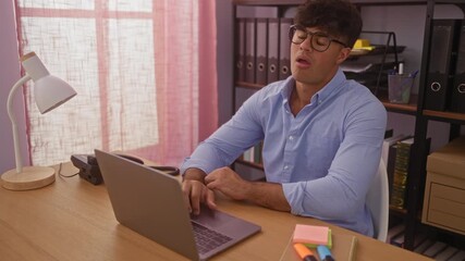 Poster - Young hispanic man working in an office sitting at a desk with a laptop, surrounded by pink curtains, lamp, and office supplies, expressing frustration.