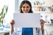 © Tapanut - Smiling female doctor in a lab coat holding a blank white sign in a medical office setting.