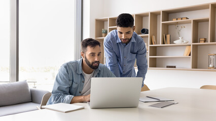 Two multiethnic company male employees talking at laptop, discussing online project, web design, application, software, looking at display, watching, reading content, working on teamwork task together