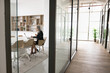 © fizkes - Mature business leader woman working at laptop in meeting room alone, sitting at large table with computer behind workspace glass wall. Contemporary office space interior with empty hall. Wide shot